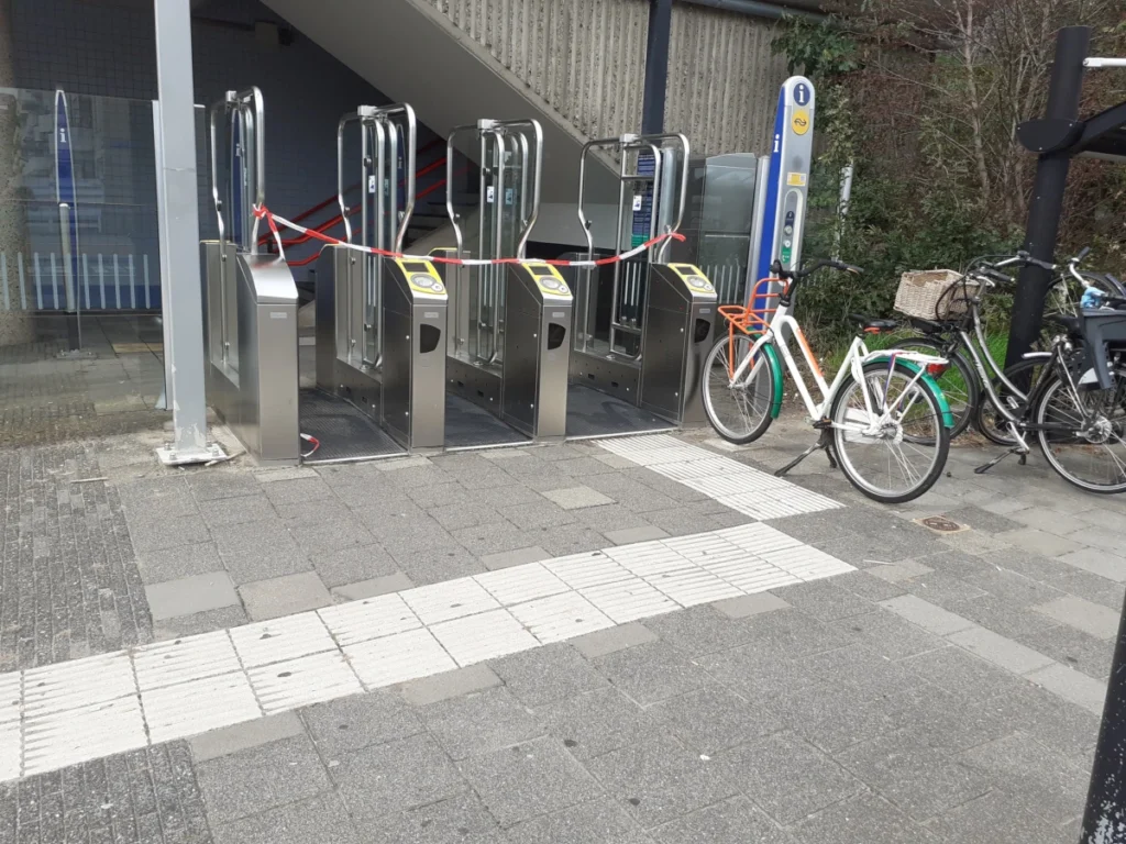 Drie toegangspoortjes op station Almere Muziekwijk. Er hangt een rood-wit lint voor de poortjes, zodat reizigers er niet doorheen kunnen. Rechts van de poortjes staan een paar fietsen geparkeerd, waaronder een witte fiets met een oranje rekje aan de voorkant. Op de grond voor de poortjes liggen tegels en een witte gidslijn voor blinden.