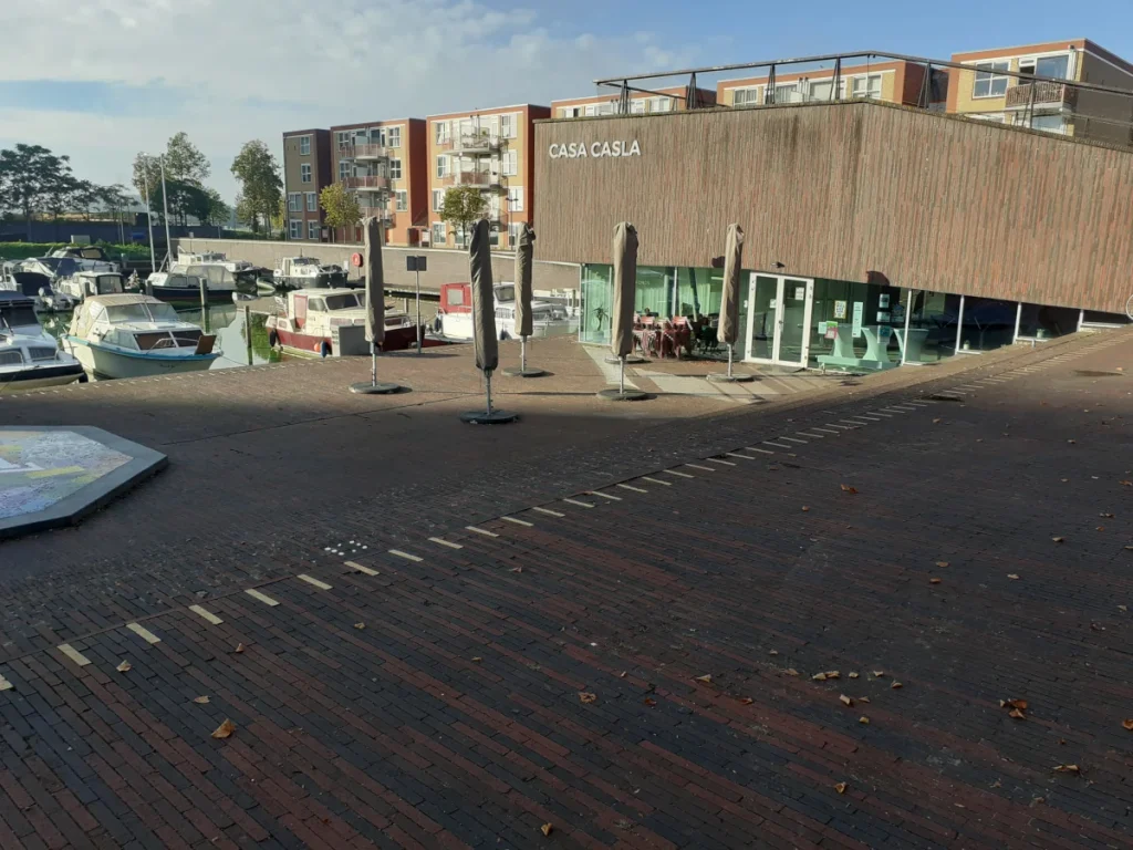 Modern houten gebouw 'Casa Casla' aan een haven met boten. Voor het gebouw ligt een terras en een plein met rode klinkers.
